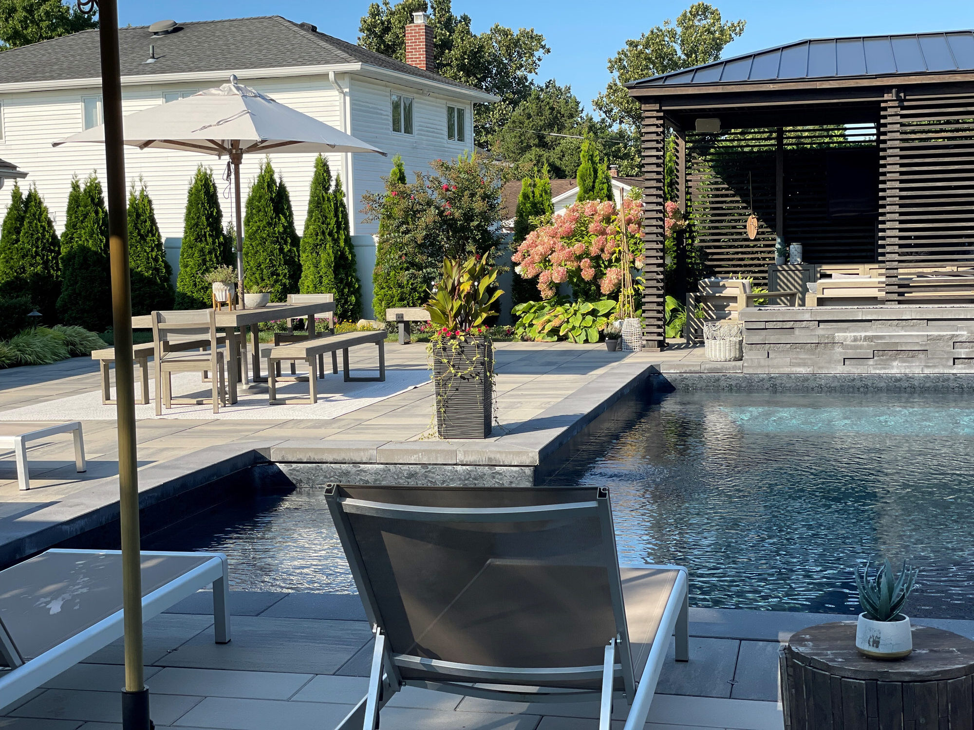 High-end, NY outdoor living space, a backyard patio with pergola, pavilion, cabana, chaise lounge chair, outdoor rug,  tequila bar, dining set with table and chairs, couch, television, pool with waterfall, umbrella, fire pit and stunning grey patio. Modern vibe. Surrounded by lush cottage style gardens,  planters, annuals, perennial flowers, shrubs and trees.  Stunning entertainment space to be used all year round and to enjoy in the summer time.  party, relax and unwind.  Cool off in the pool, stay warm by the fire.